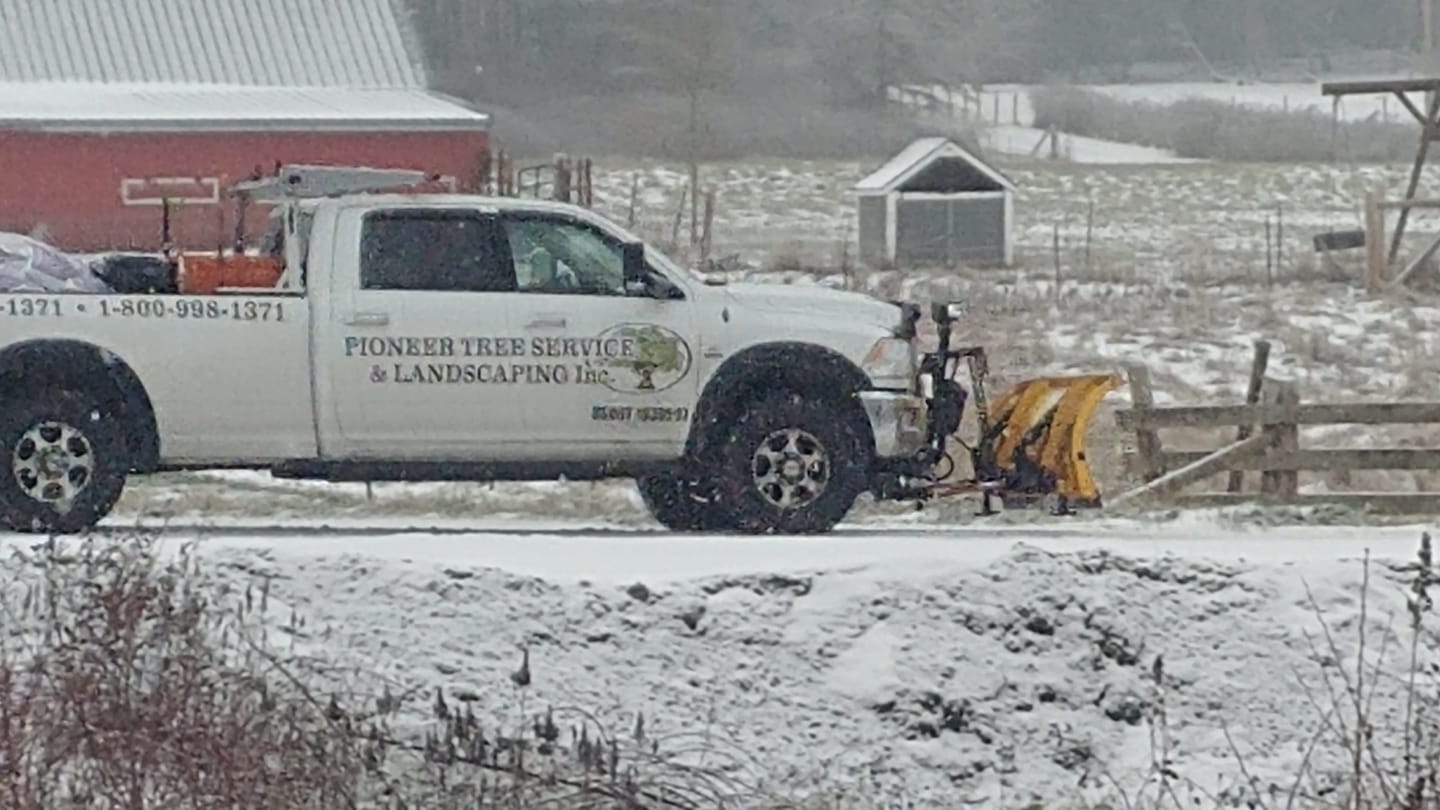 White pickup truck with snowplow clearing a snow-covered road near a red barn and fence.