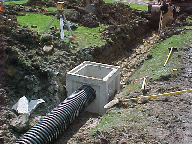 Construction site with a concrete drainage box, corrugated pipe, and trench dug in the ground.