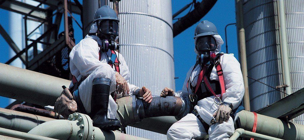 Two men wearing gas masks are sitting on a pipe.