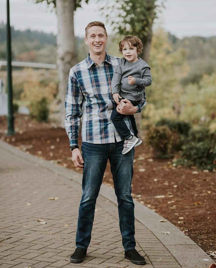 Man holding a smiling child outdoors on a curved path in plaid shirts and jeans, with trees in the background