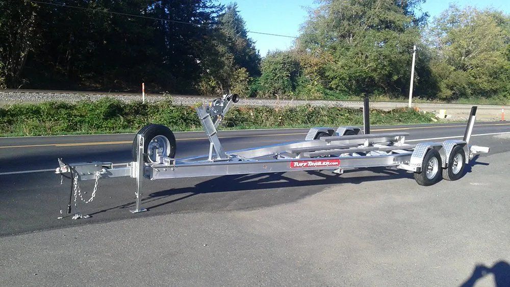 Silver boat trailer parked on asphalt road. Spare tire on side, supports and rollers visible.