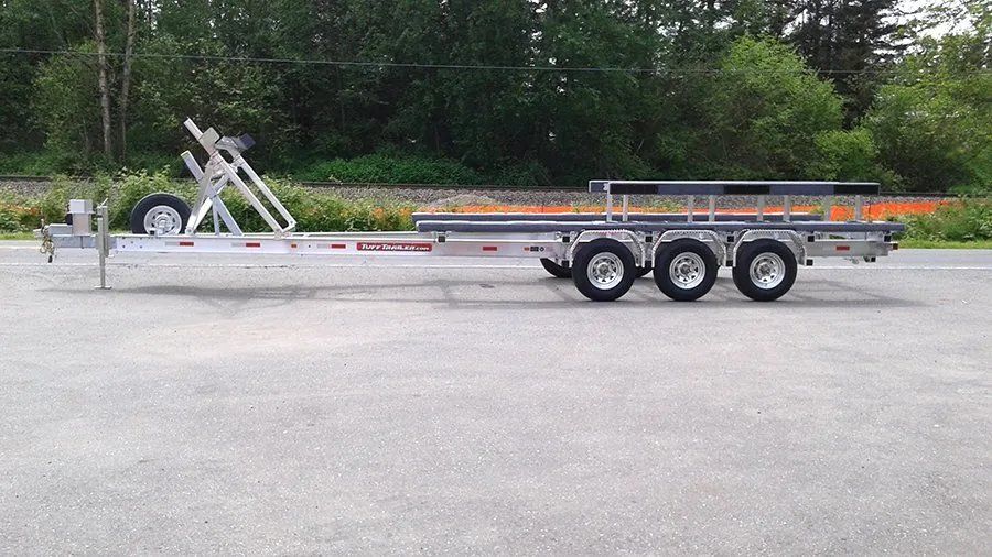 Silver boat trailer on pavement, three axles, winch on front, green trees in background.