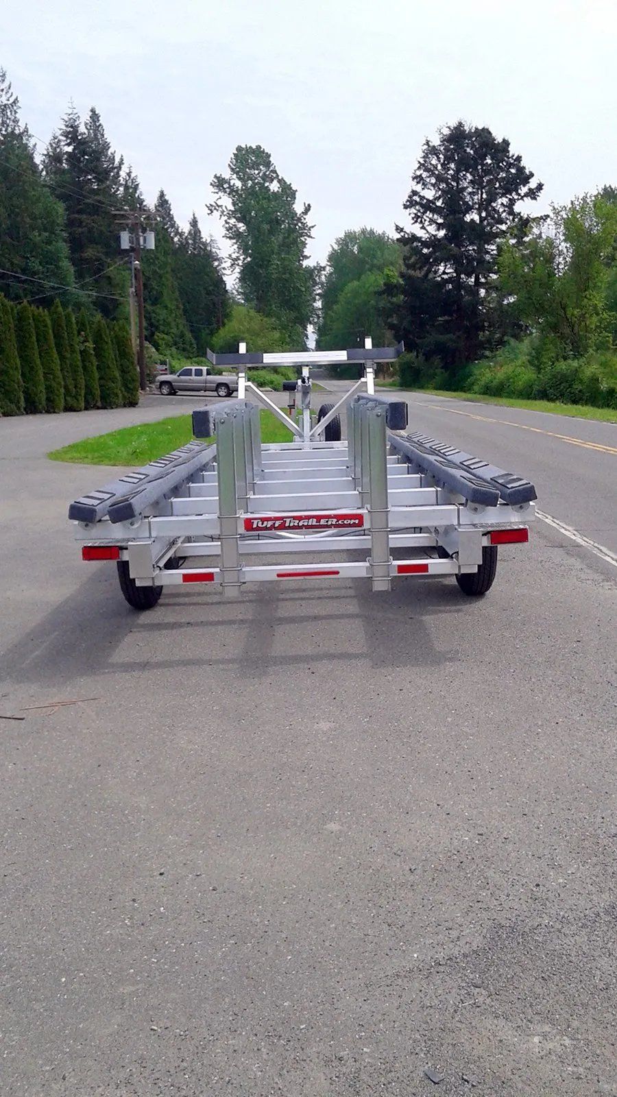 Boat trailer parked on a paved road, silver metal frame, rollers, red reflectors, trees in the background.