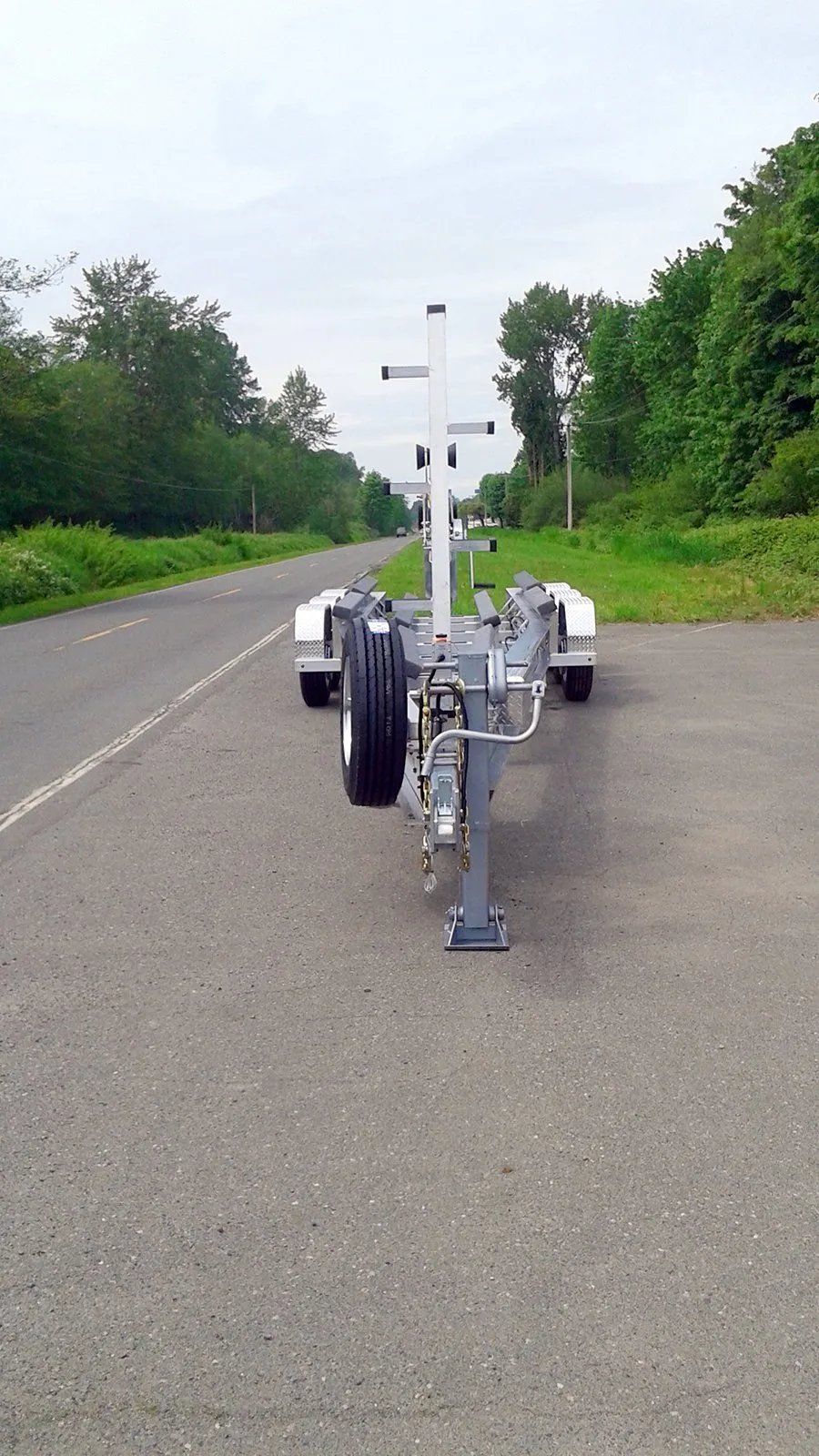 Boat trailer parked on asphalt road, spare tire, white vertical frame, trees in the background.