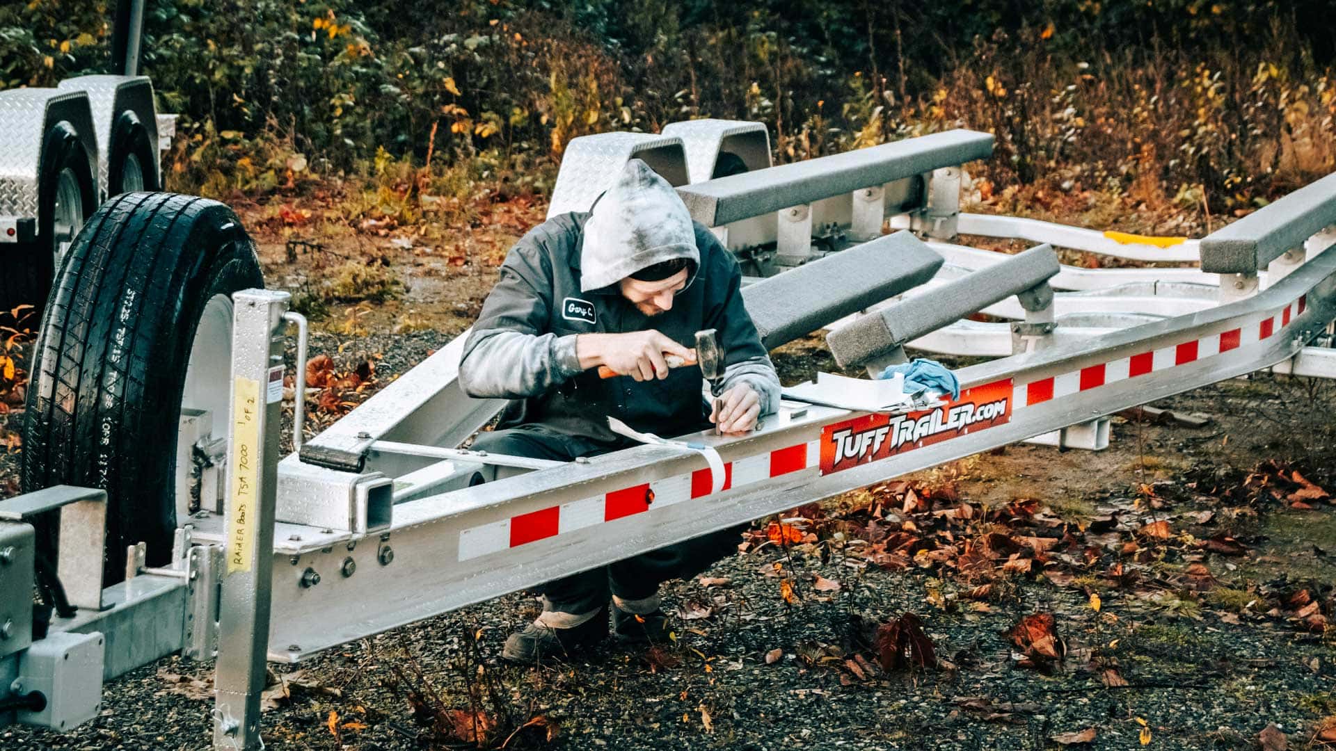 Person in hoodie working on a boat trailer with a hammer outdoors.