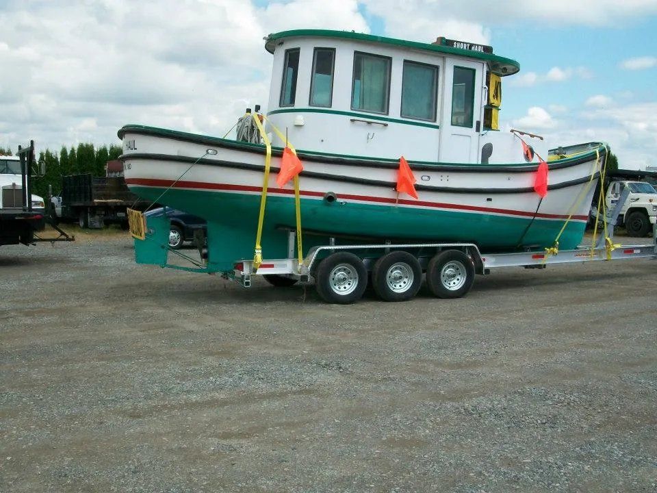 Green and white tugboat on a trailer, secured with yellow straps, parked on gravel.