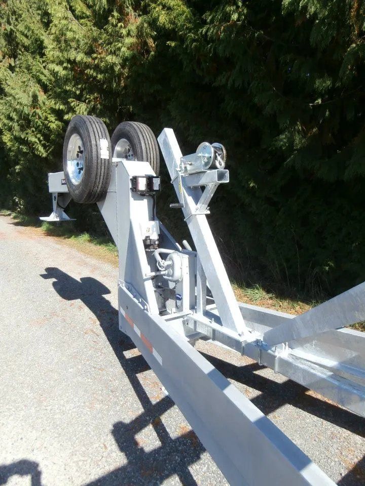 A gray boat trailer angled on pavement, with a spare tire, winch, and supports. Green trees in background.
