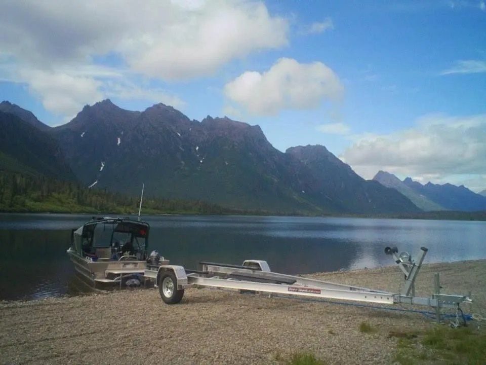 Boat on a trailer at a lake shore with mountain backdrop, under a partly cloudy sky.
