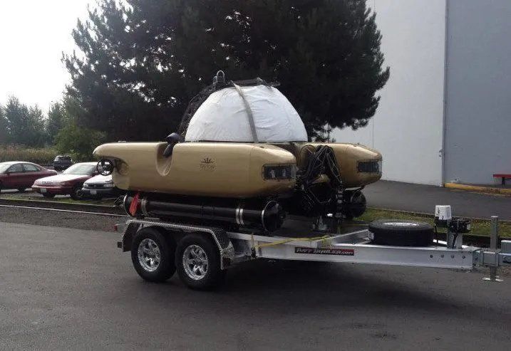A yellow submersible on a trailer; parked on pavement in front of a building and some cars.