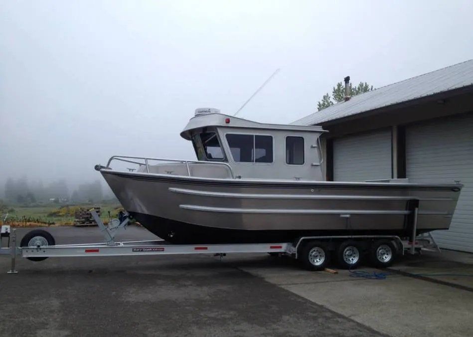 Aluminum boat on a trailer, outside, with a covered building in the background on an overcast day.