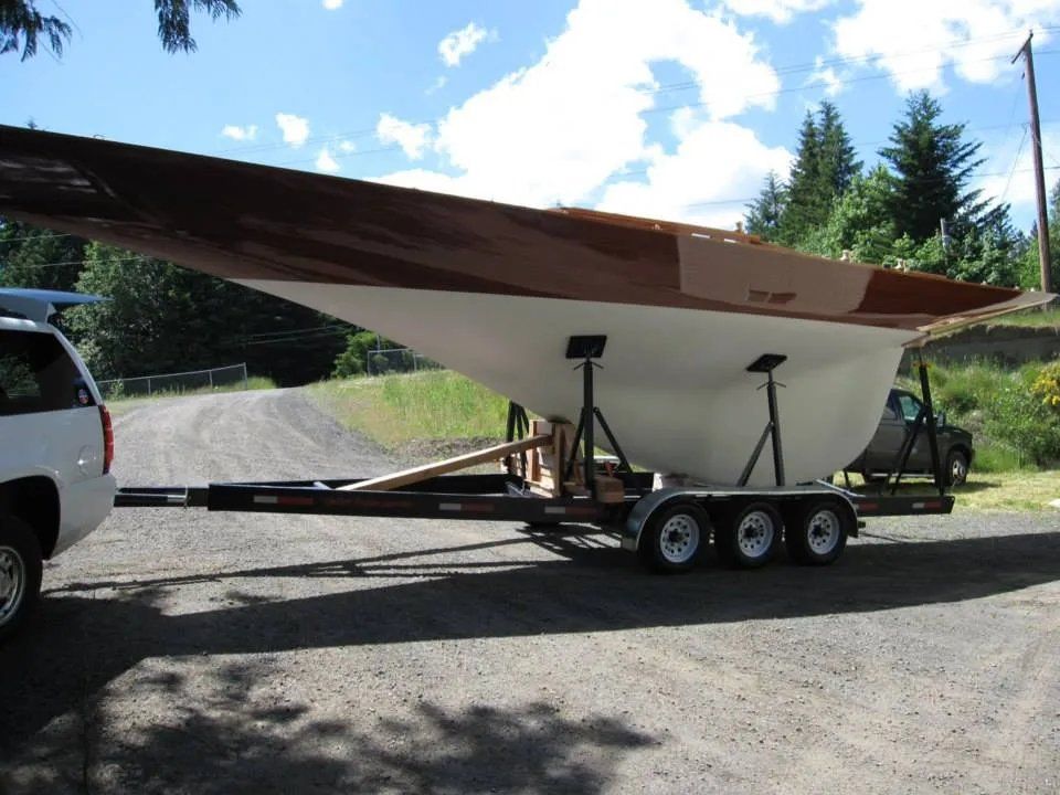 A sailboat on a trailer, with a white hull and brown deck, parked on asphalt.