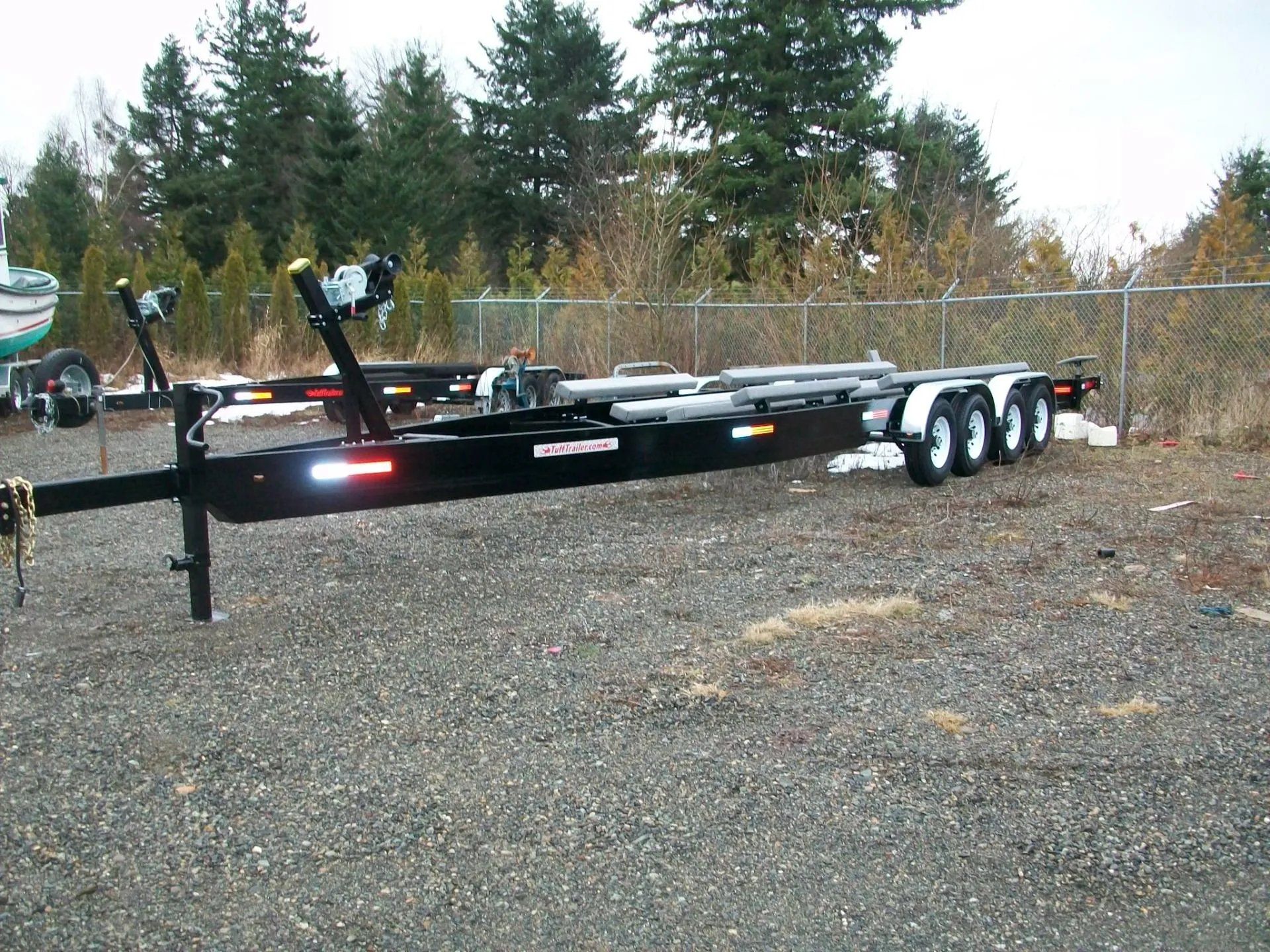 Black boat trailer parked on gravel, with three axles, lights, and rollers.