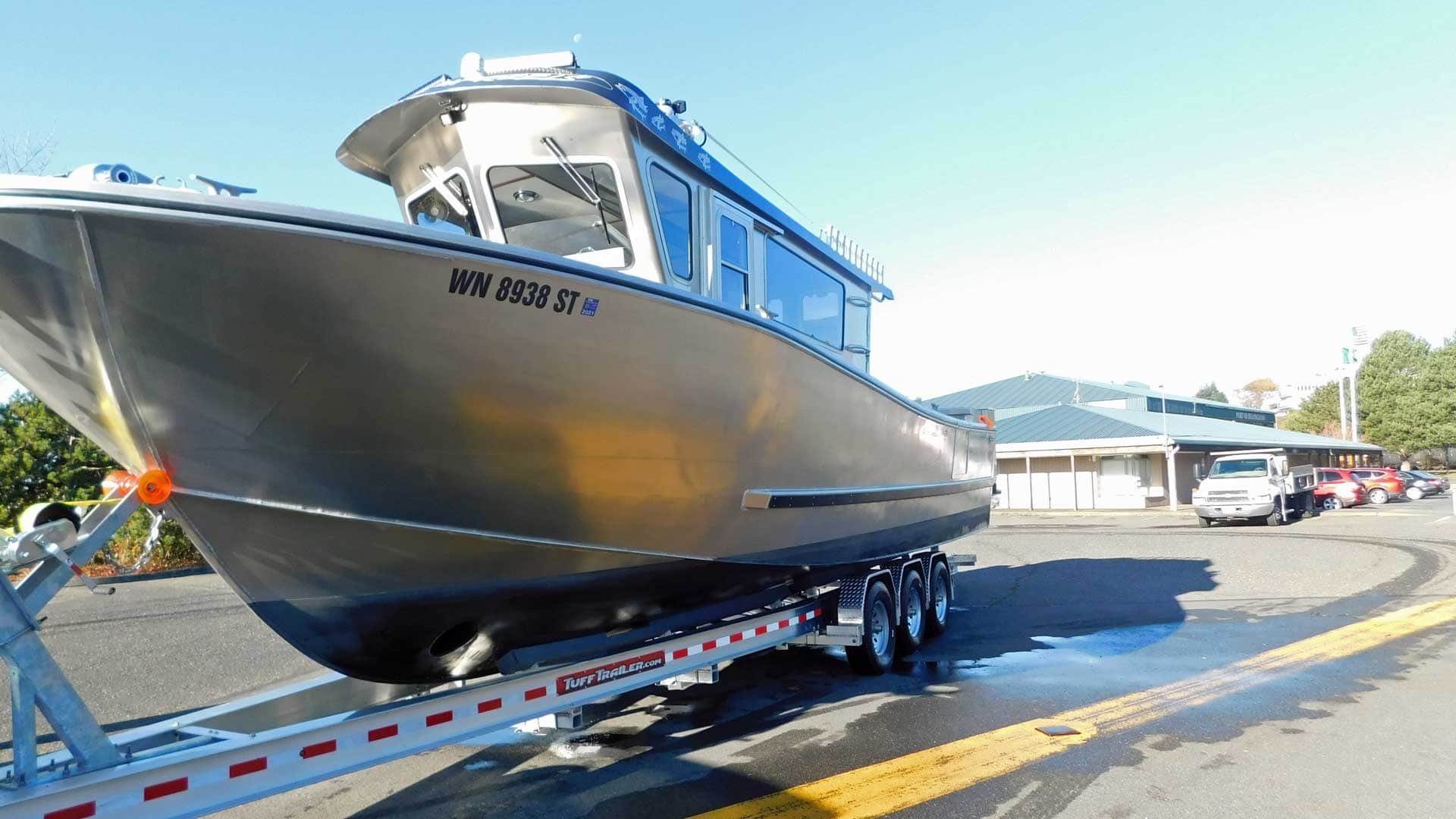A large silver boat on a trailer, parked on a road next to a building on a sunny day.