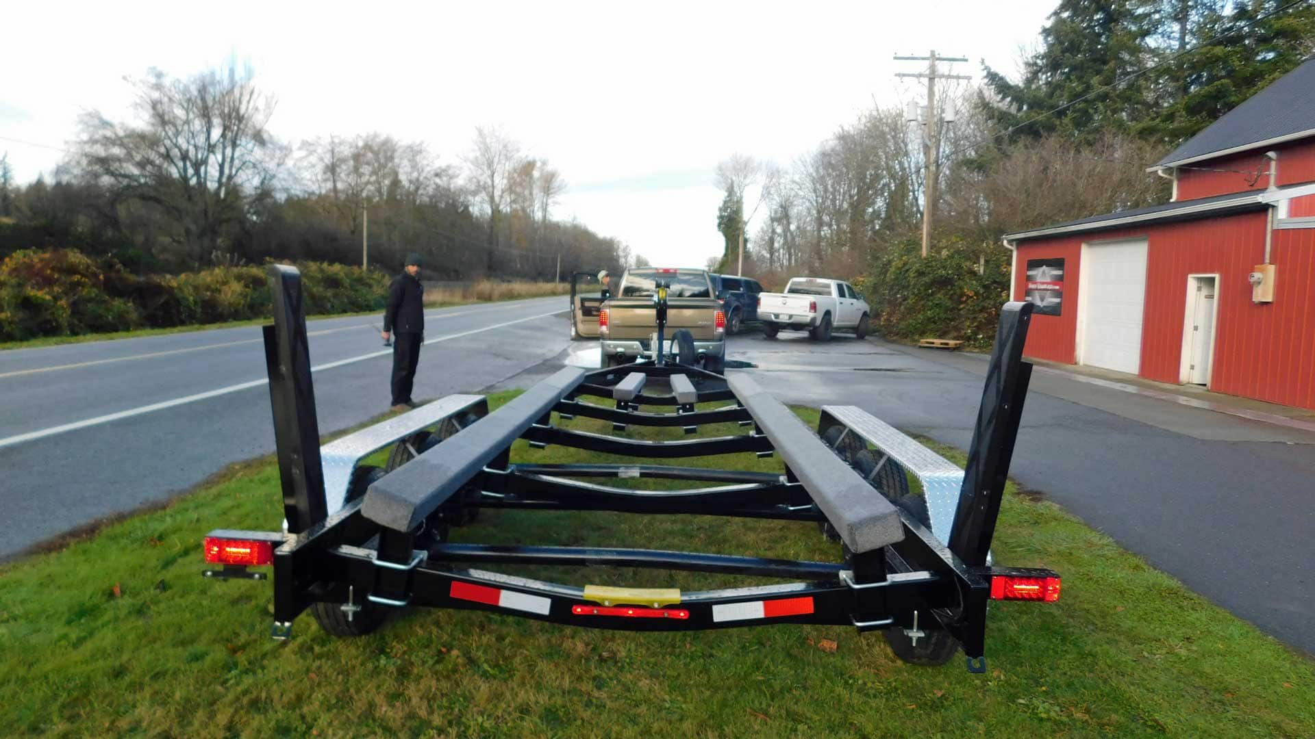 Boat trailer on grass next to a road, with cars and a person standing nearby. Red building in background.