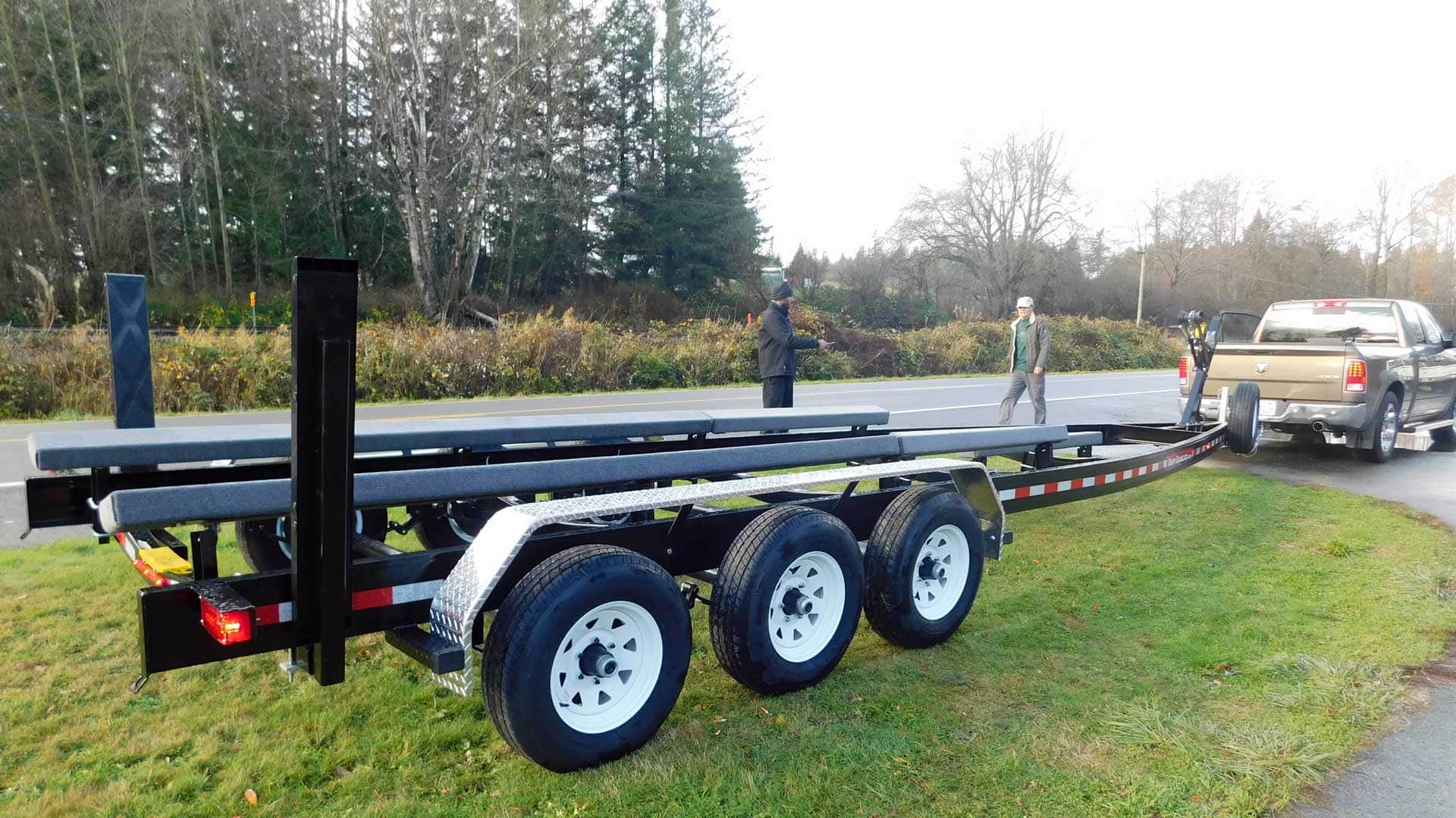 Boat trailer pulled by a truck on a grassy roadside, three people visible.