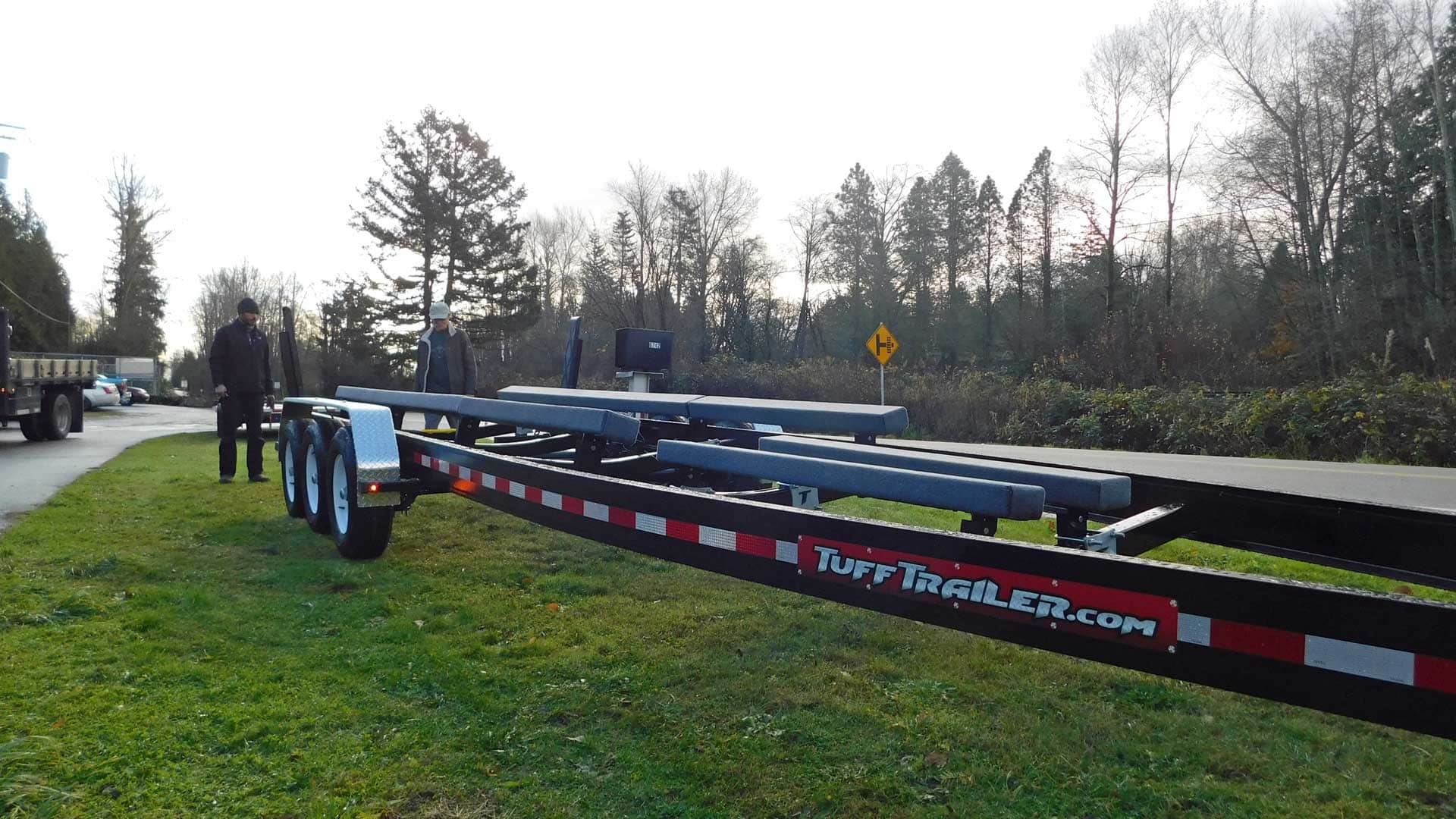 Boat trailer on grass next to a road, person standing nearby.