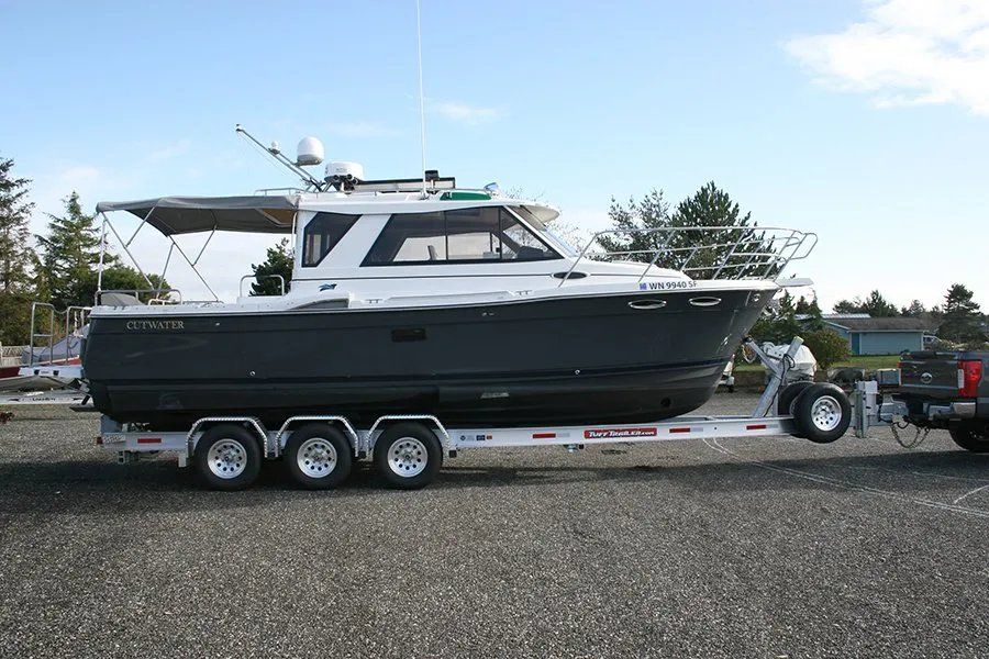 Gray boat on a trailer, parked on pavement; a truck is attached.