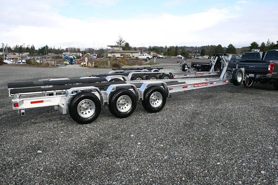 Aluminum boat trailer with three axles parked on gravel, with a truck attached.
