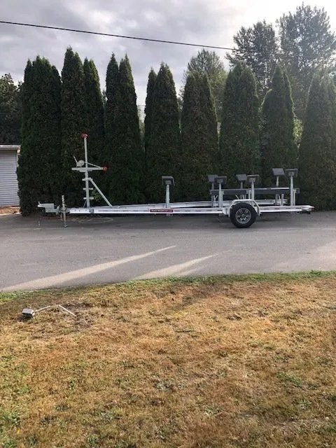 Boat trailer parked on asphalt in front of tall evergreen trees.