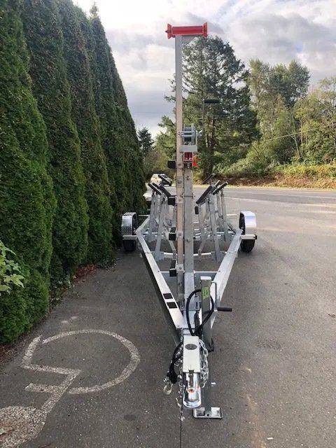 New silver boat trailer parked on asphalt in front of evergreen hedge.