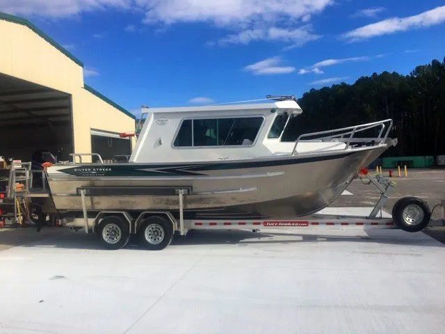 Aluminum boat on a trailer, with a white cabin, parked in front of a tan building under a blue sky.
