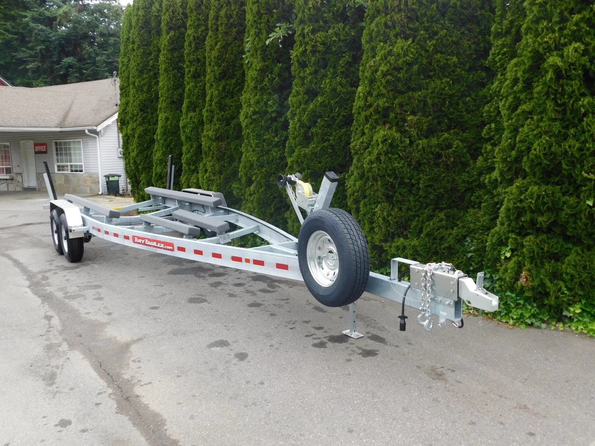 Silver boat trailer parked on a paved surface, spare tire mounted, with green hedge background.