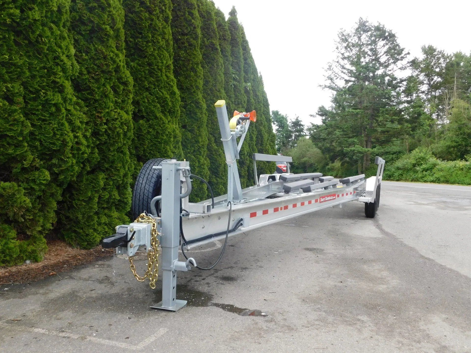 Boat trailer parked on asphalt next to a line of tall green trees.