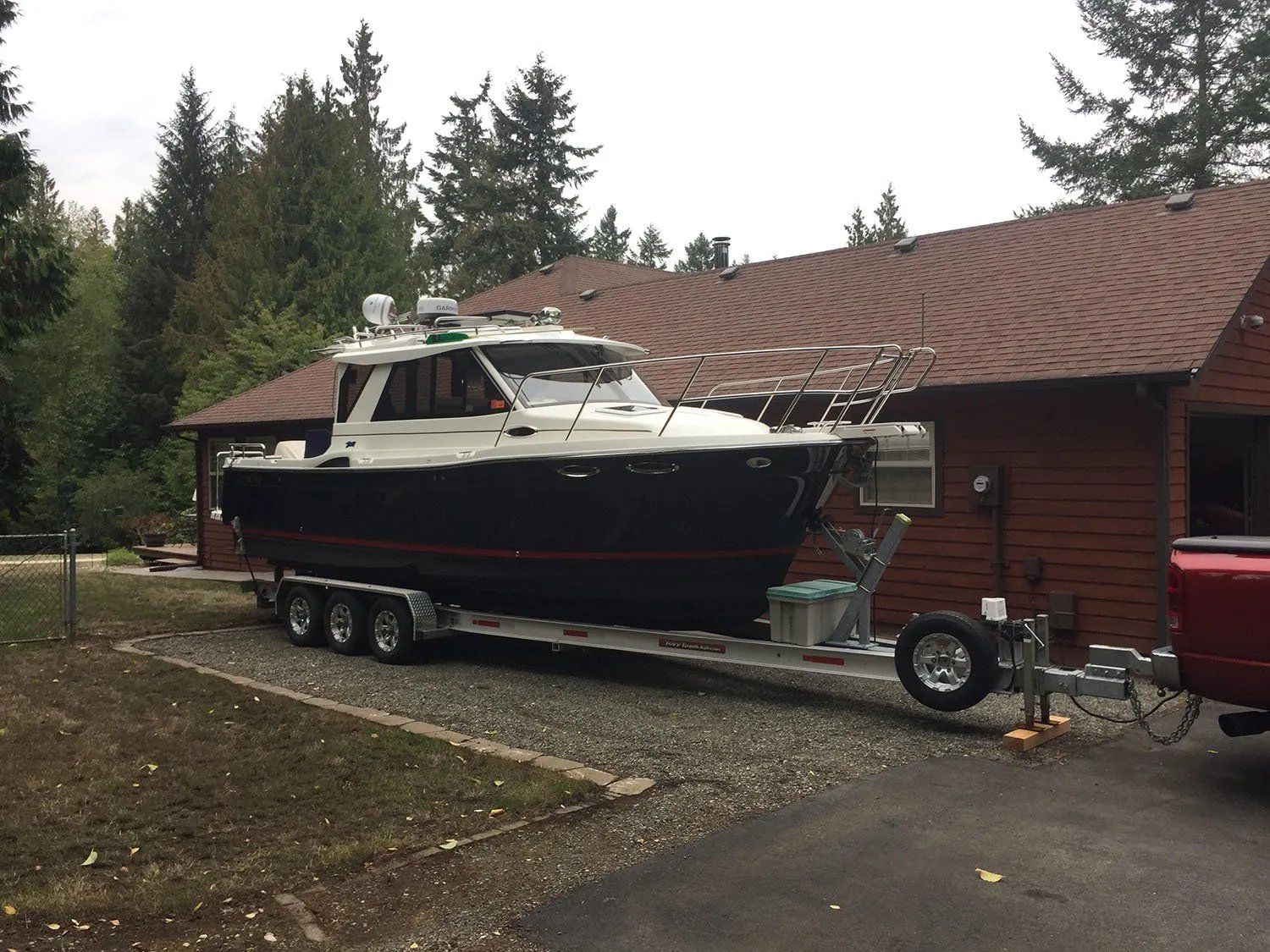 Boat on trailer in front of a house. Dark blue hull, white top, brown roof, and green trees in background.
