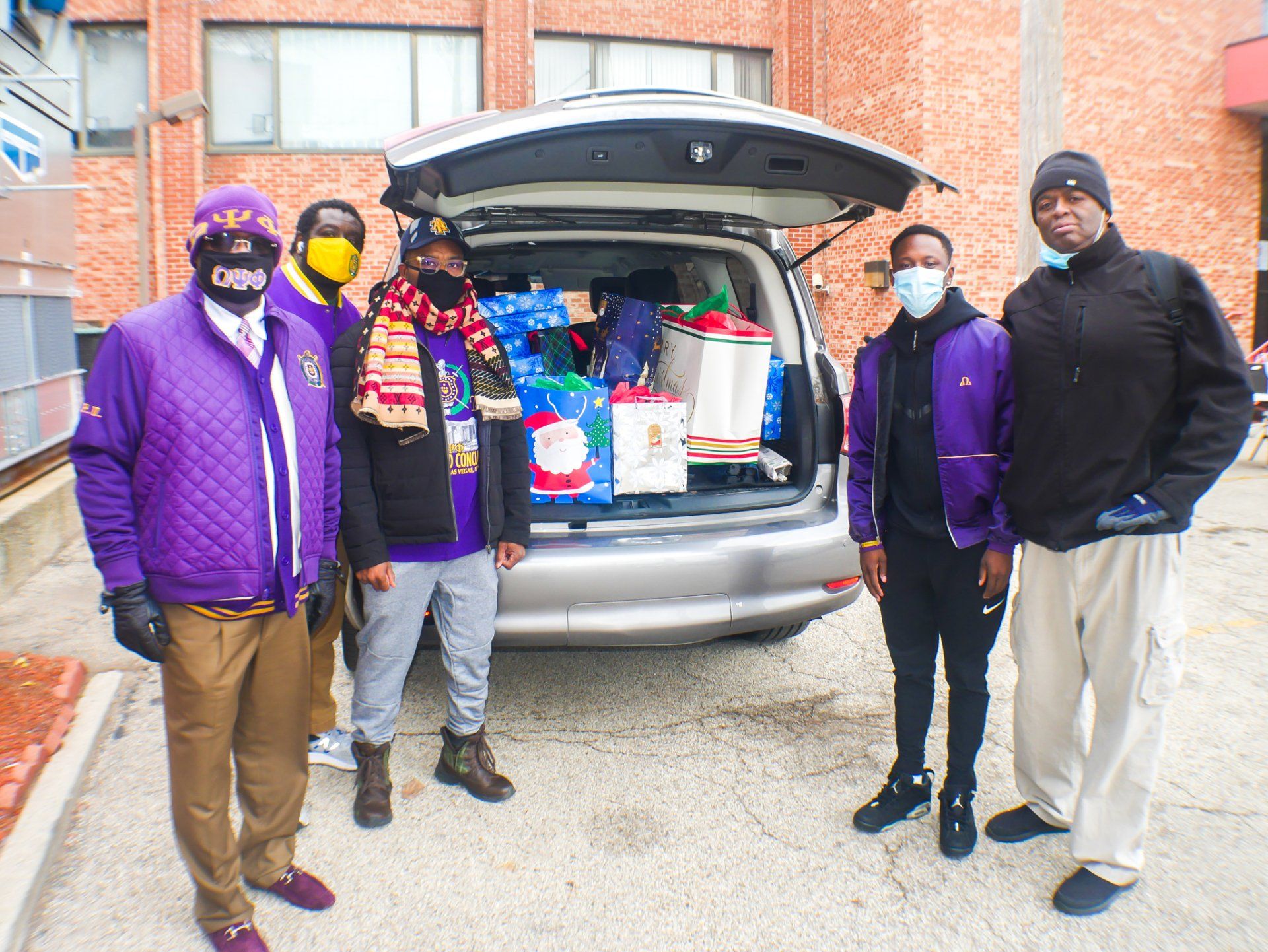 A group of people wearing masks are standing in front of a van filled with gifts.