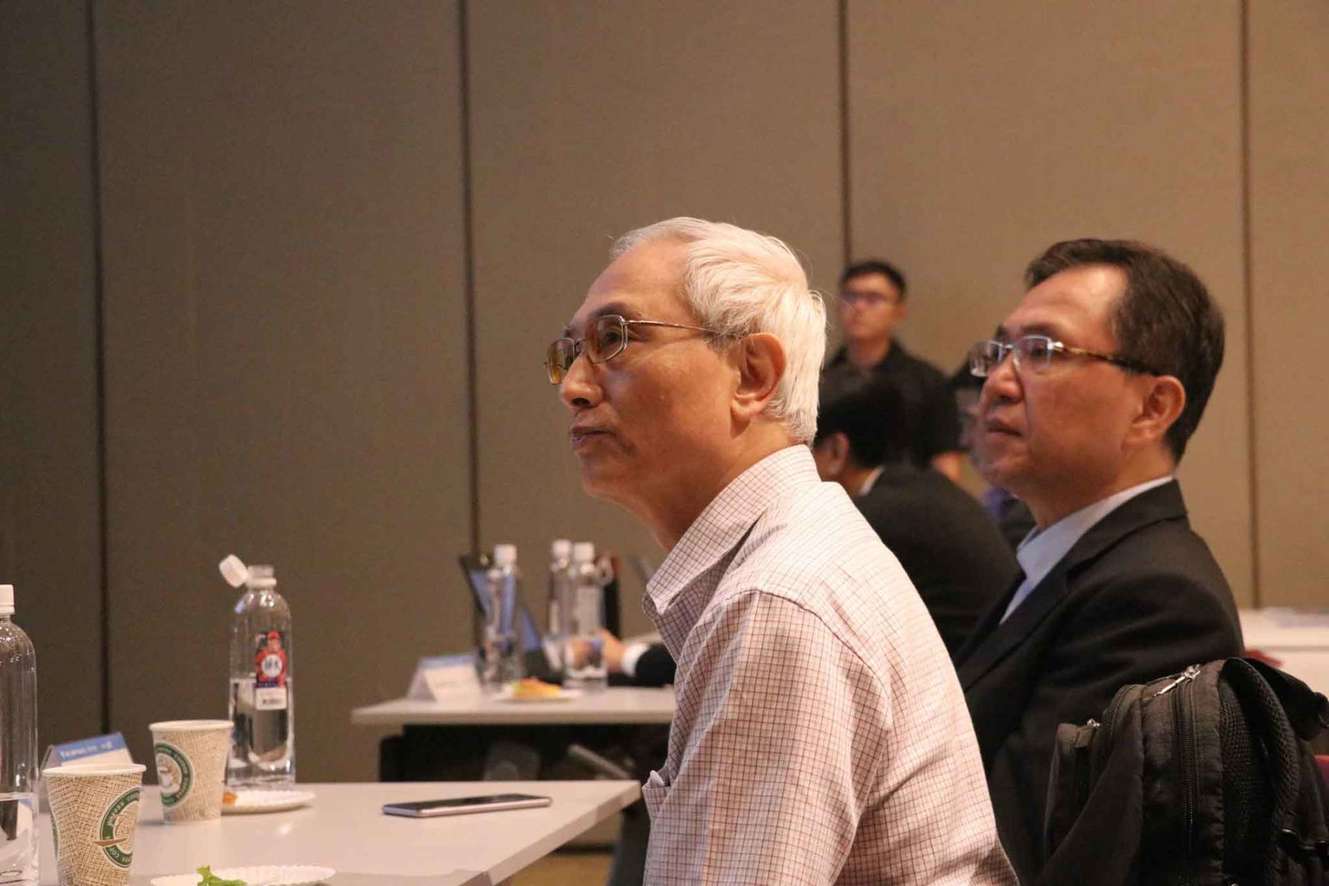 會議室裡，兩名男子認真地聽著。一名穿著淺色襯衫，一名穿著西裝。