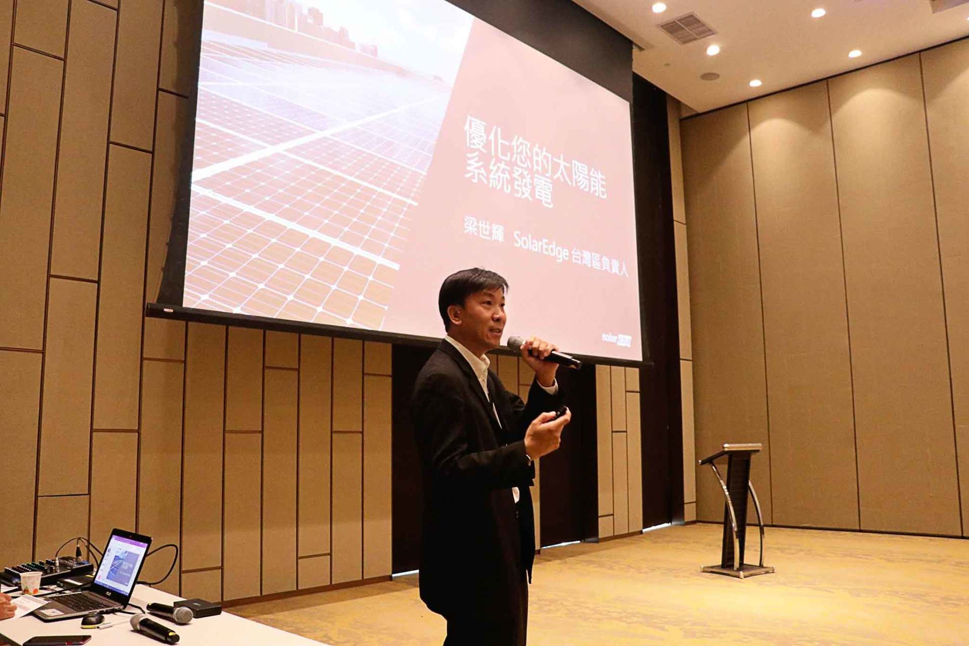 一名男子在會議上發言，手持麥克風。房間為米色，螢幕上顯示文字和太陽能板。