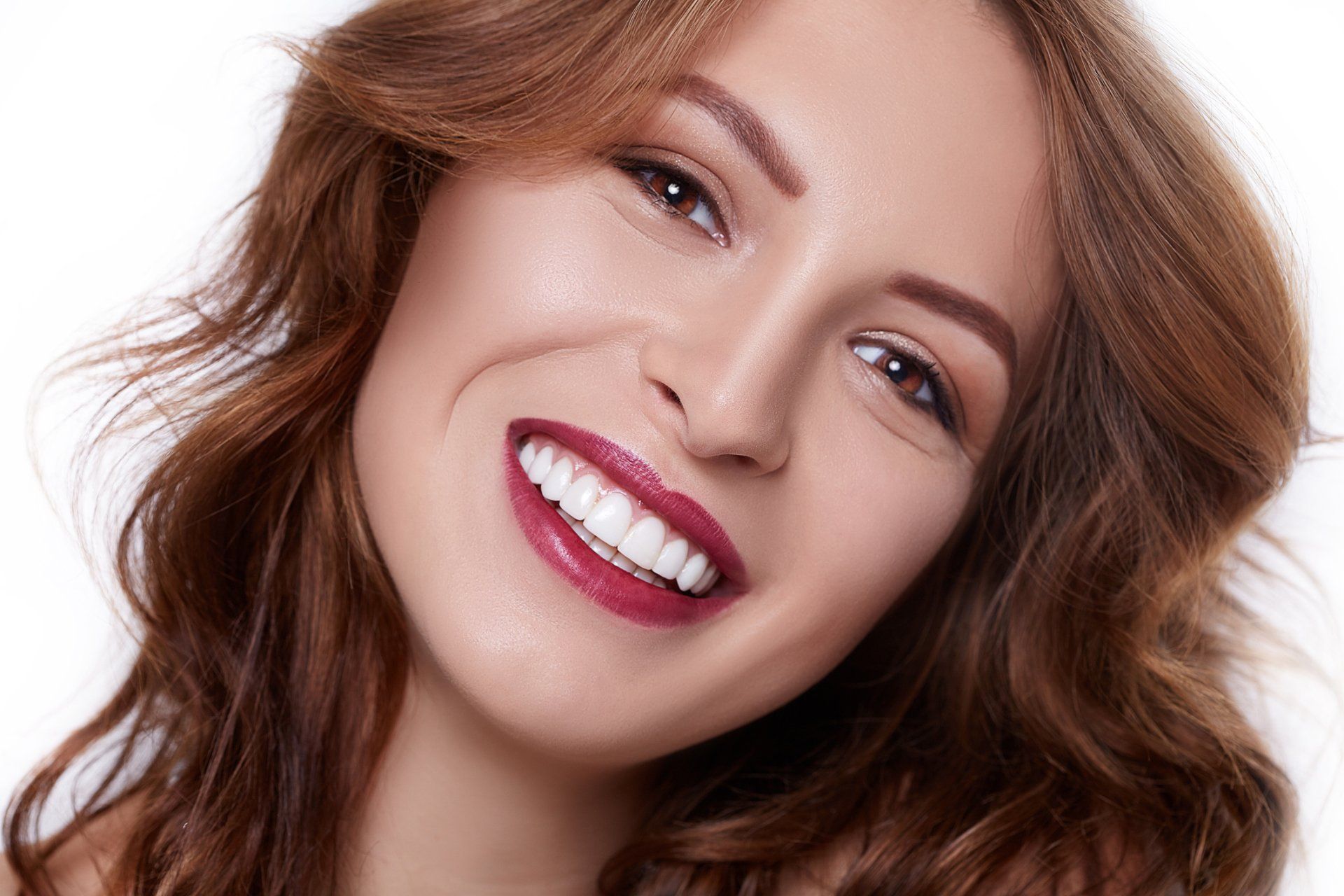 Woman with wavy brown hair smiles widely, showing white teeth and dark red lipstick.