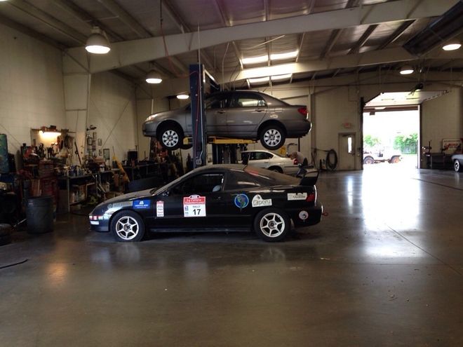 A black sports car and a silver car on a lift inside a garage with open door.