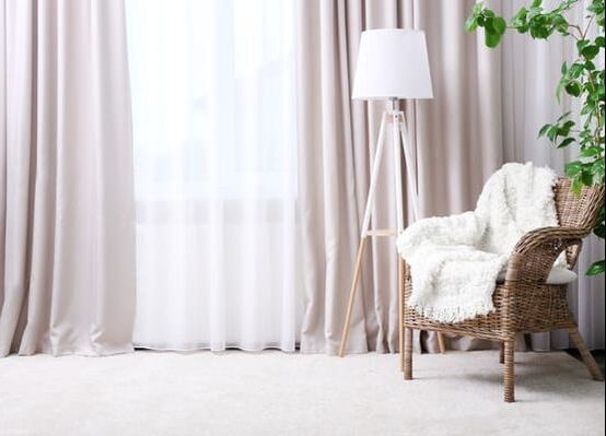 Soft pink curtains over sheet white curtains, behind a cane chair, with lamp and green plant.