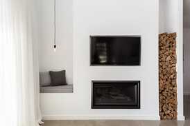 White indoor room with sheer white curtains. Facing wall with built in fireplace, wall mounted TV, wood stacked into recess in the wall.