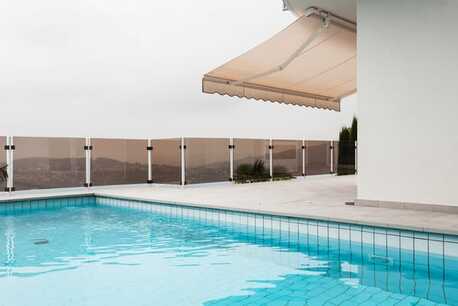 Outdoor awning extending out over pool paving, with pool in foreground, and looking out over pool fence at mountain terrain in background.