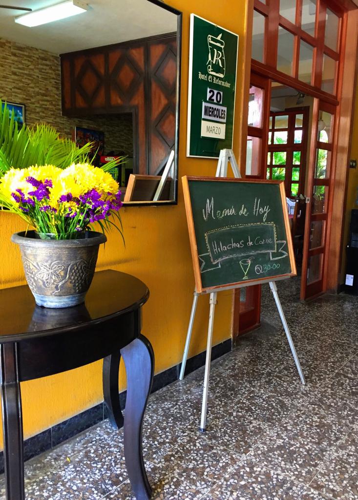 Entrada de la cafetería con un letrero de pizarra, un jarrón con flores moradas y amarillas y una puerta de madera 