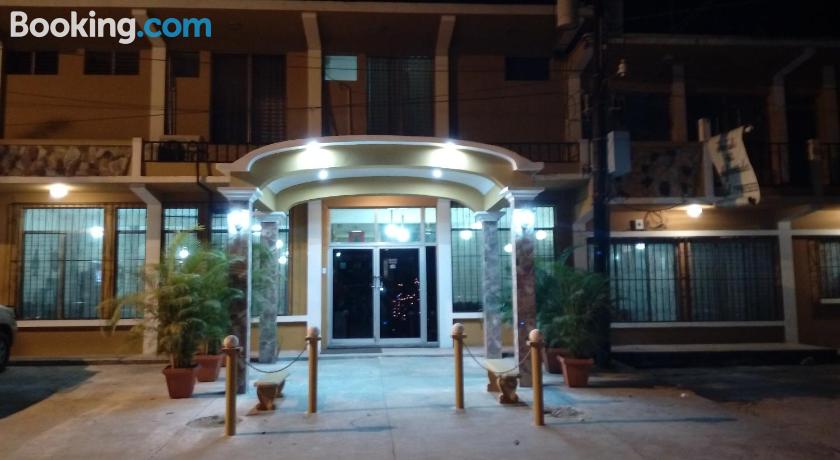 Vista nocturna de la entrada de un hotel con un toldo iluminado, puertas de cristal y plantas en macetas.