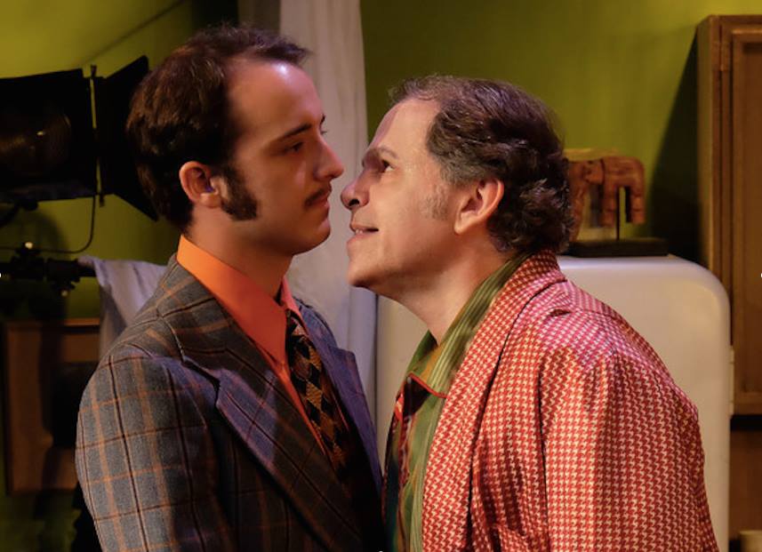 Man with sideburns and orange shirt with man in red checked jacket next to him