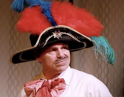 1 actor onstage wearing black hat with red and blue feathers, white shirt and scarf tie
