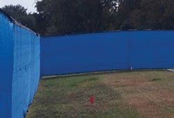 Blue outfield fence in a baseball field, green grass, and trees in the background.