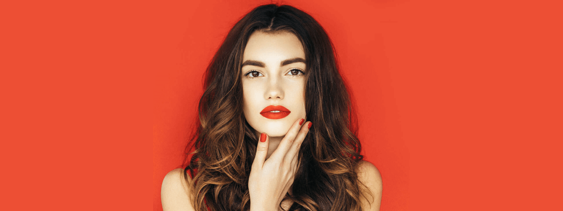 Woman with red lips and nails against a red background, touching her face.