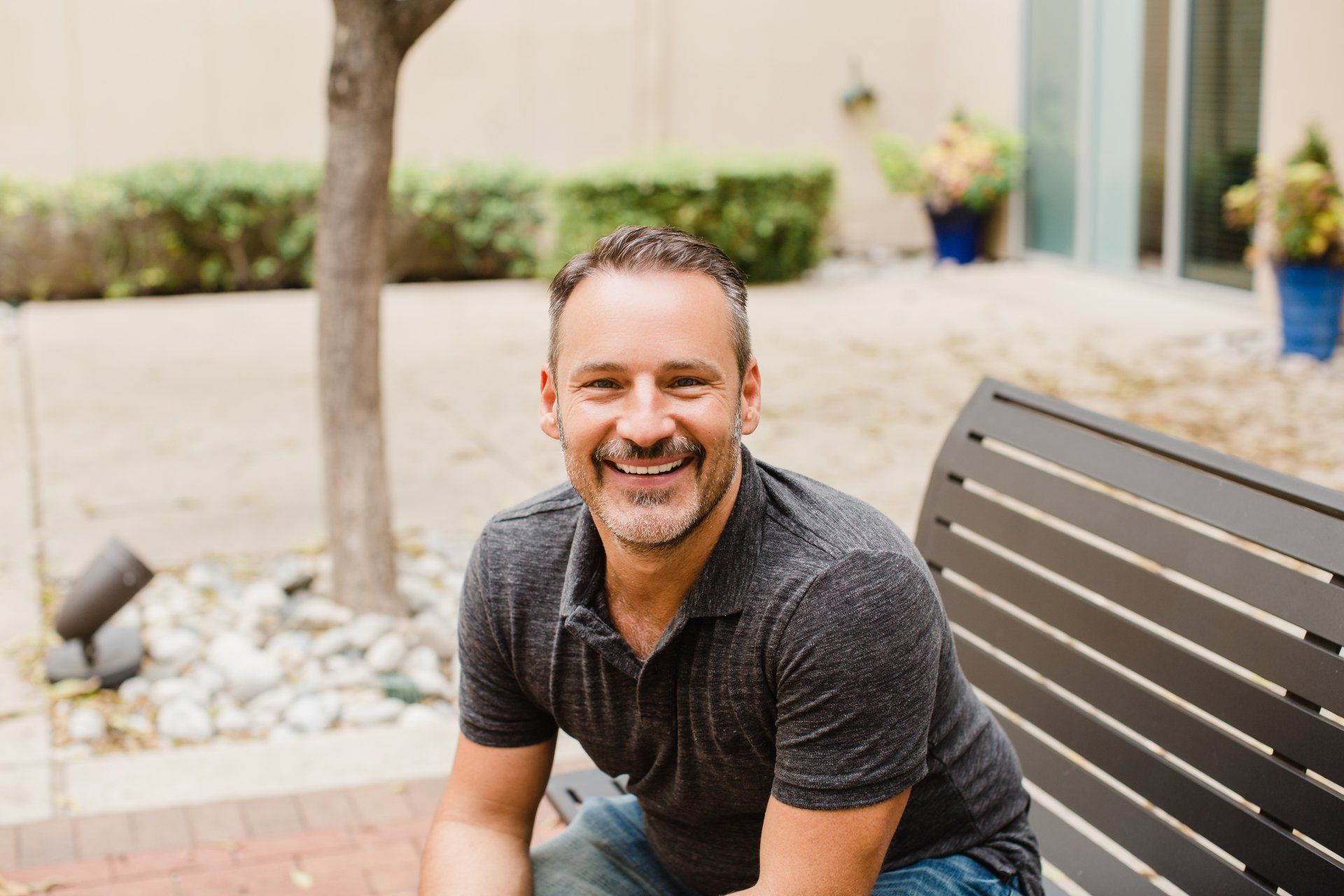 Man smiling, sitting on a bench outdoors. Gray shirt, trees and plants in the background.