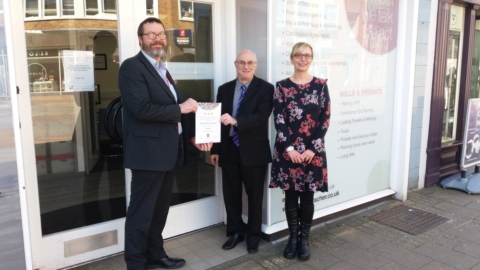 Three people are standing in front of a building holding a certificate.