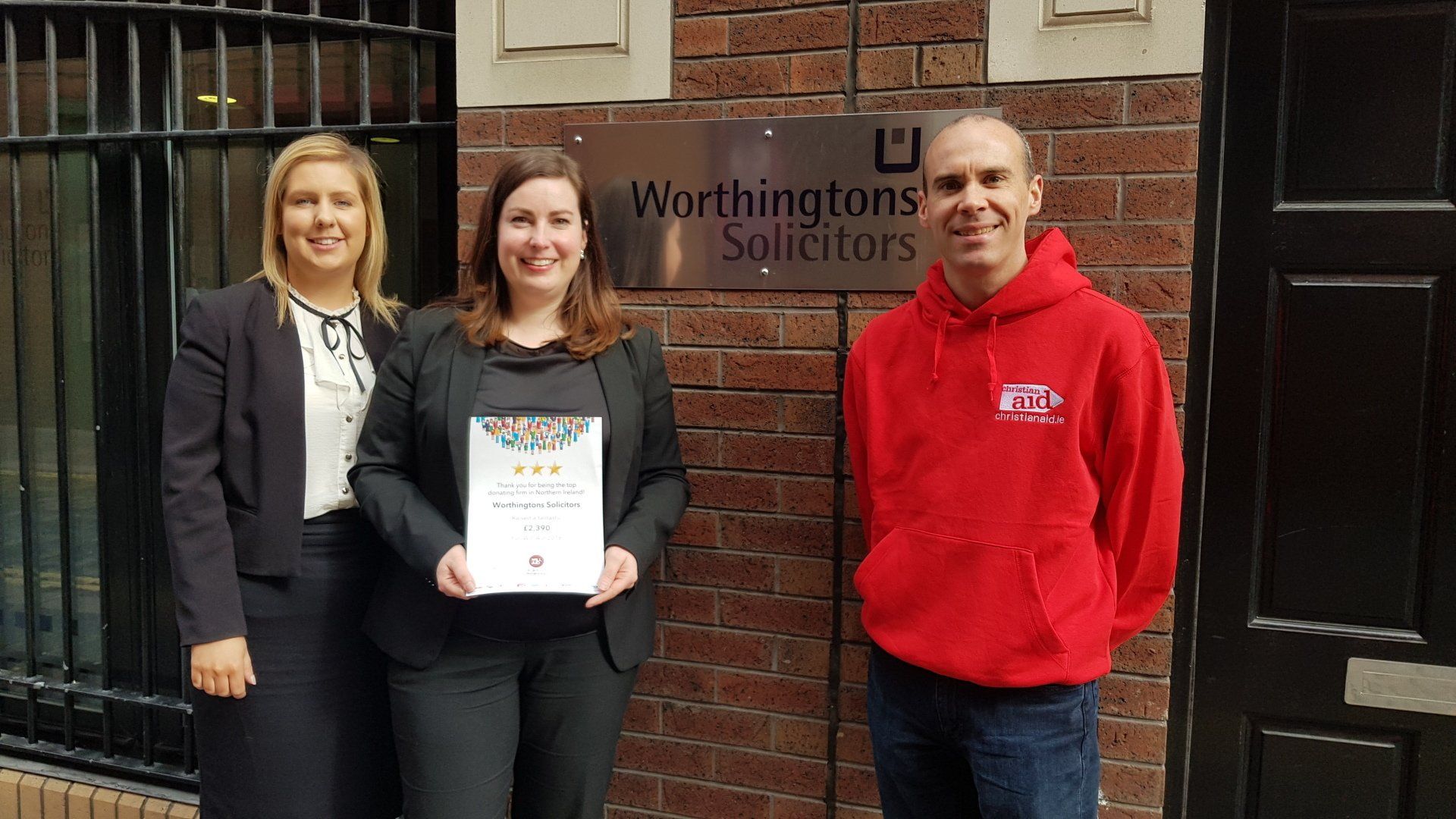 Three people are standing in front of a brick building holding a certificate.