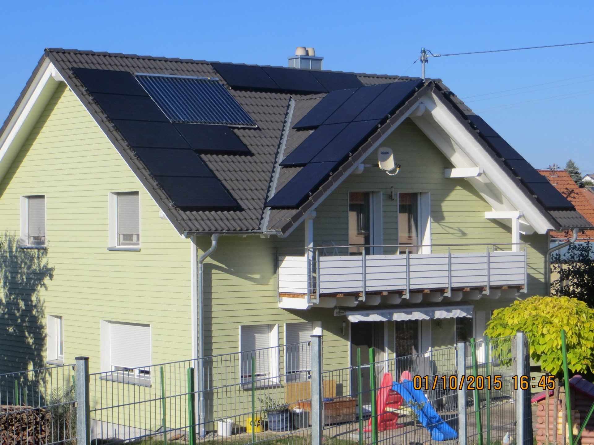 Haus mit Solarmodulen auf dem Dach, grüner Außenverkleidung, weißen Zierleisten und einem Balkon.