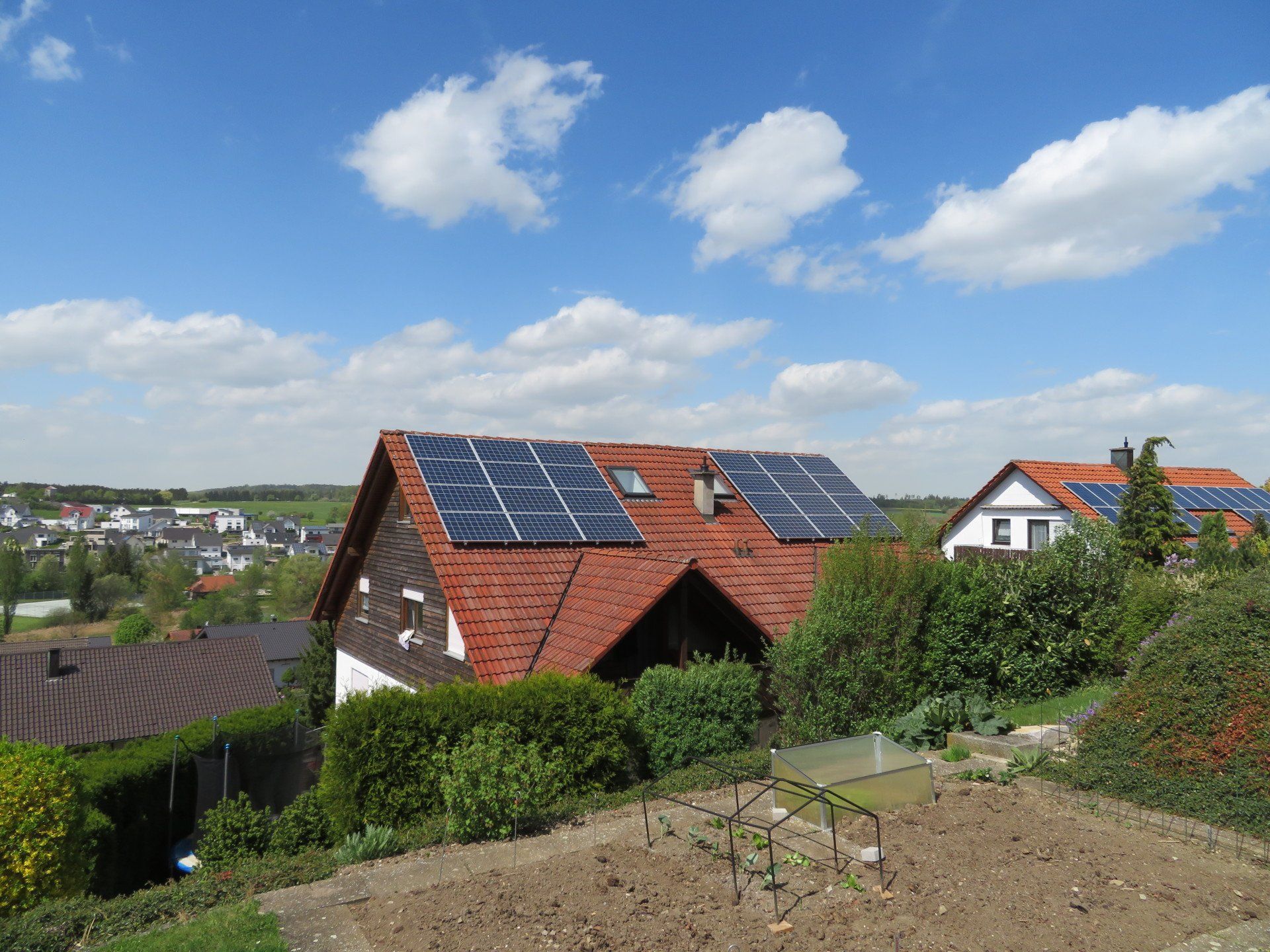 Ein Haus mit Solarzellen auf dem roten Dach, unter einem blauen Himmel mit Wolken, in einer grünen Landschaft.