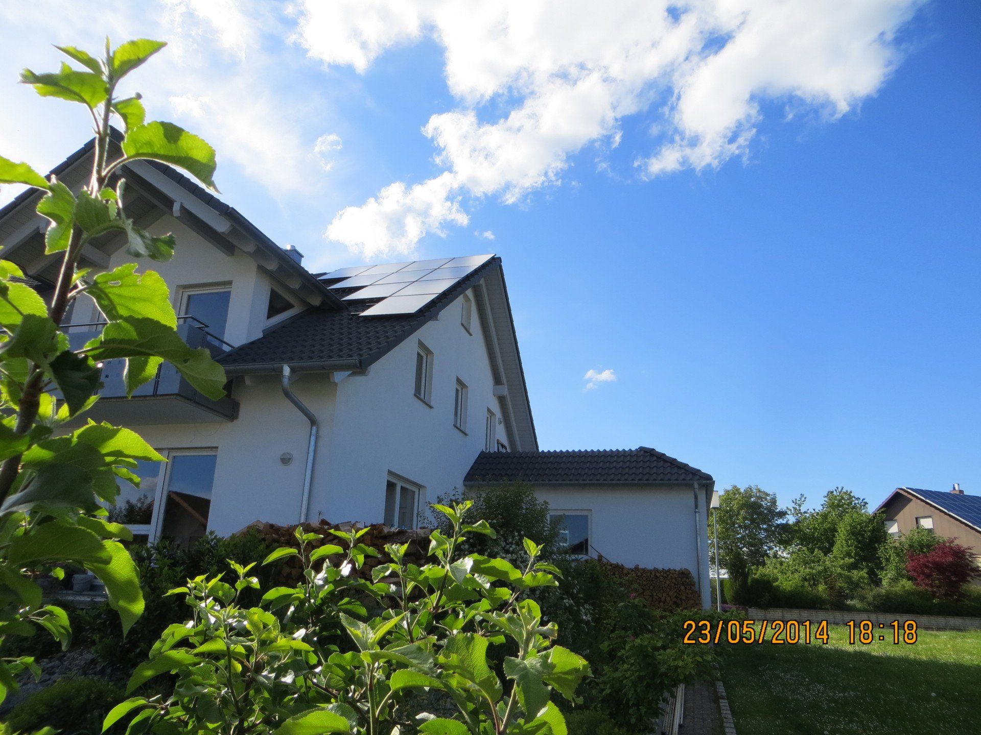 Weißes Haus mit Solarzellen, grünem Laub, blauem Himmel und einigen Wolken.