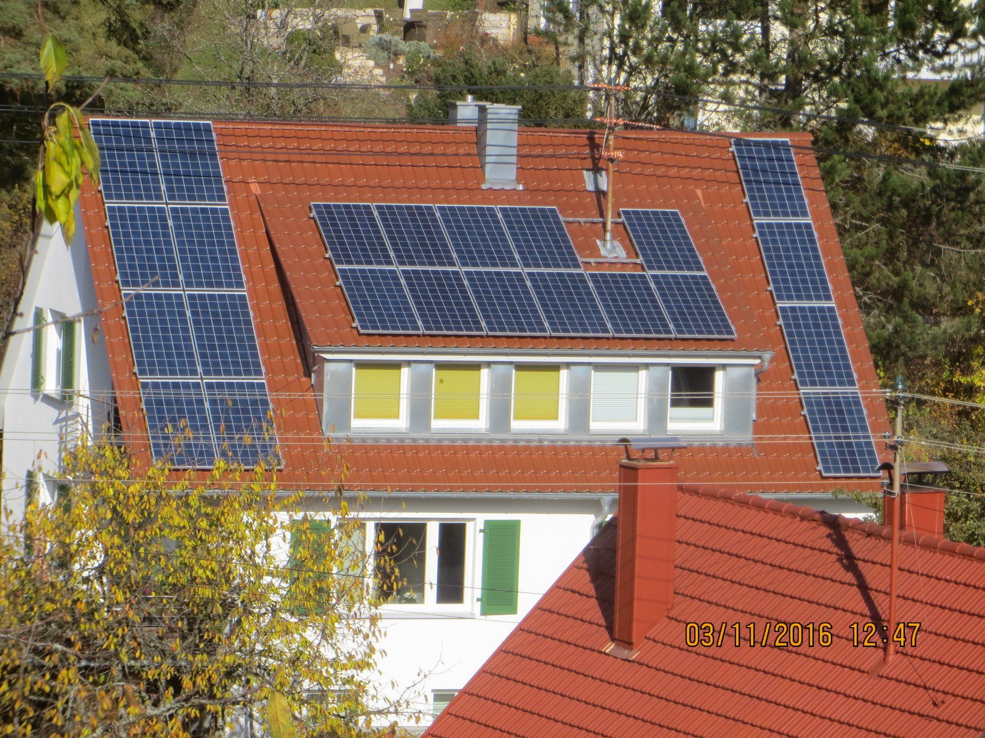 Haus mit rotem Ziegeldach und Solarmodulen. Grüne Fensterläden, gelbe Jalousien und Bäume im Hintergrund.