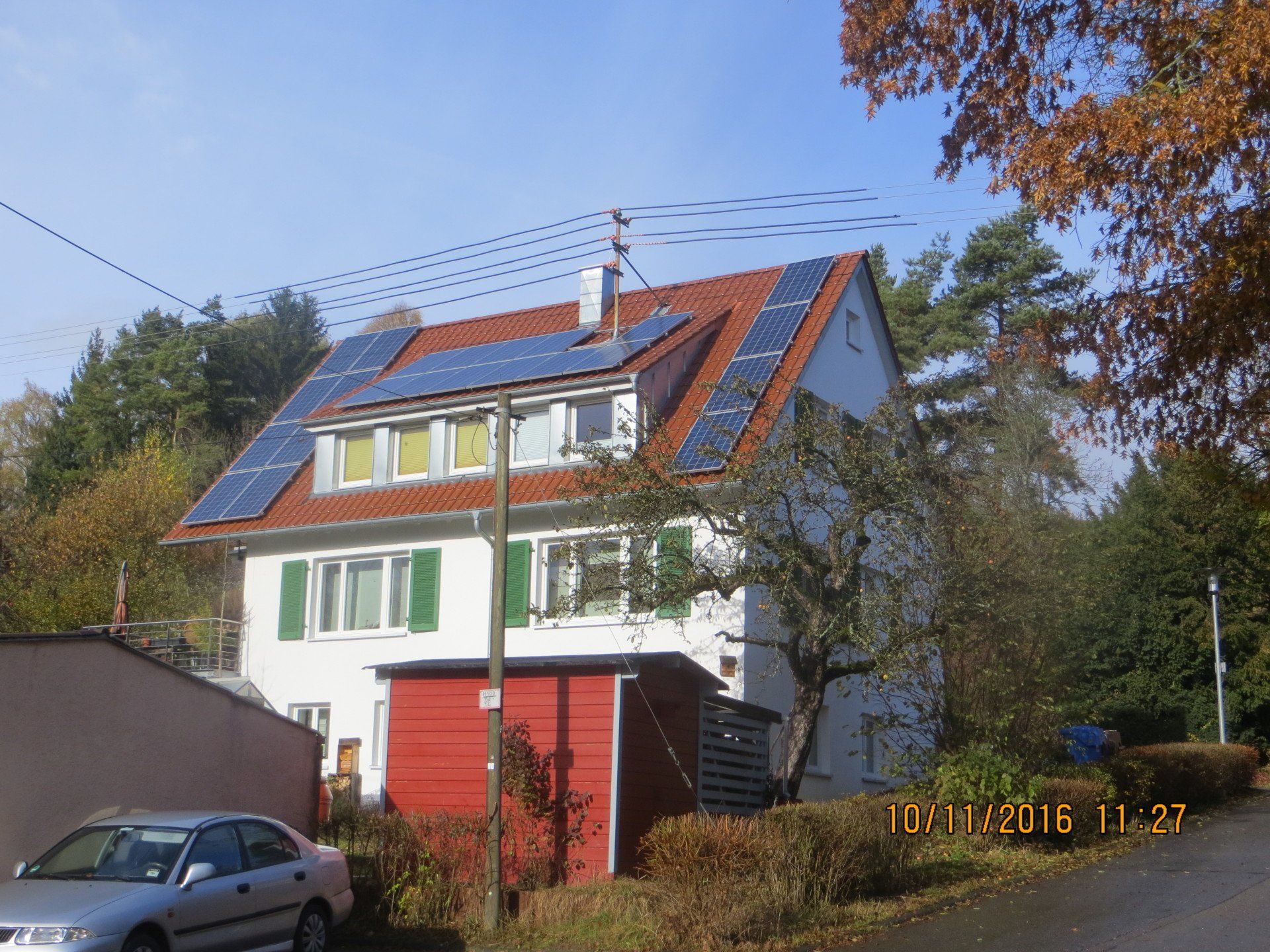 Weißes Haus mit rotem Dach, Sonnenkollektoren, grünen Fensterläden und Herbstbäumen an einem sonnigen Tag.